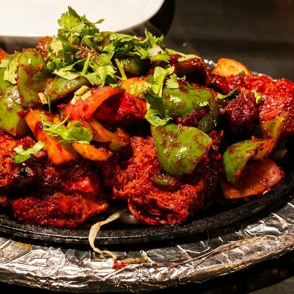 Sizzling red chicken tikka with green bell peppers, onions, and cilantro on a hot plate.